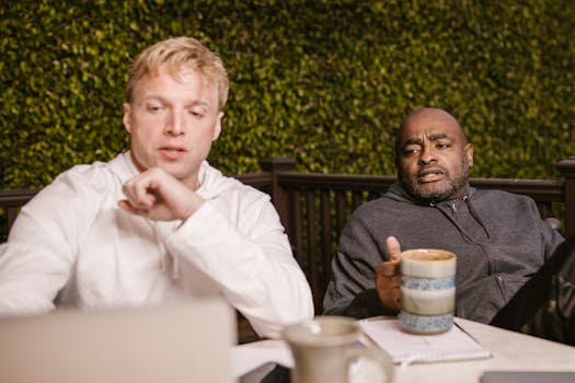 Two adults collaborating outdoors with coffee mugs, discussing work on a laptop.