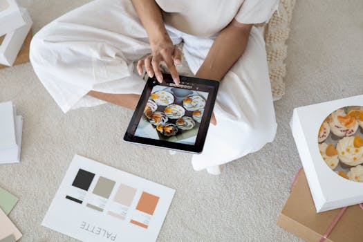 An individual selects cupcakes via tablet, surrounded by palette and cupcake box indoors.