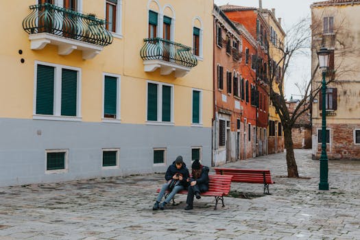 Two people seated on a bench in a vibrant Italian street, focusing on their phones on a calm day.